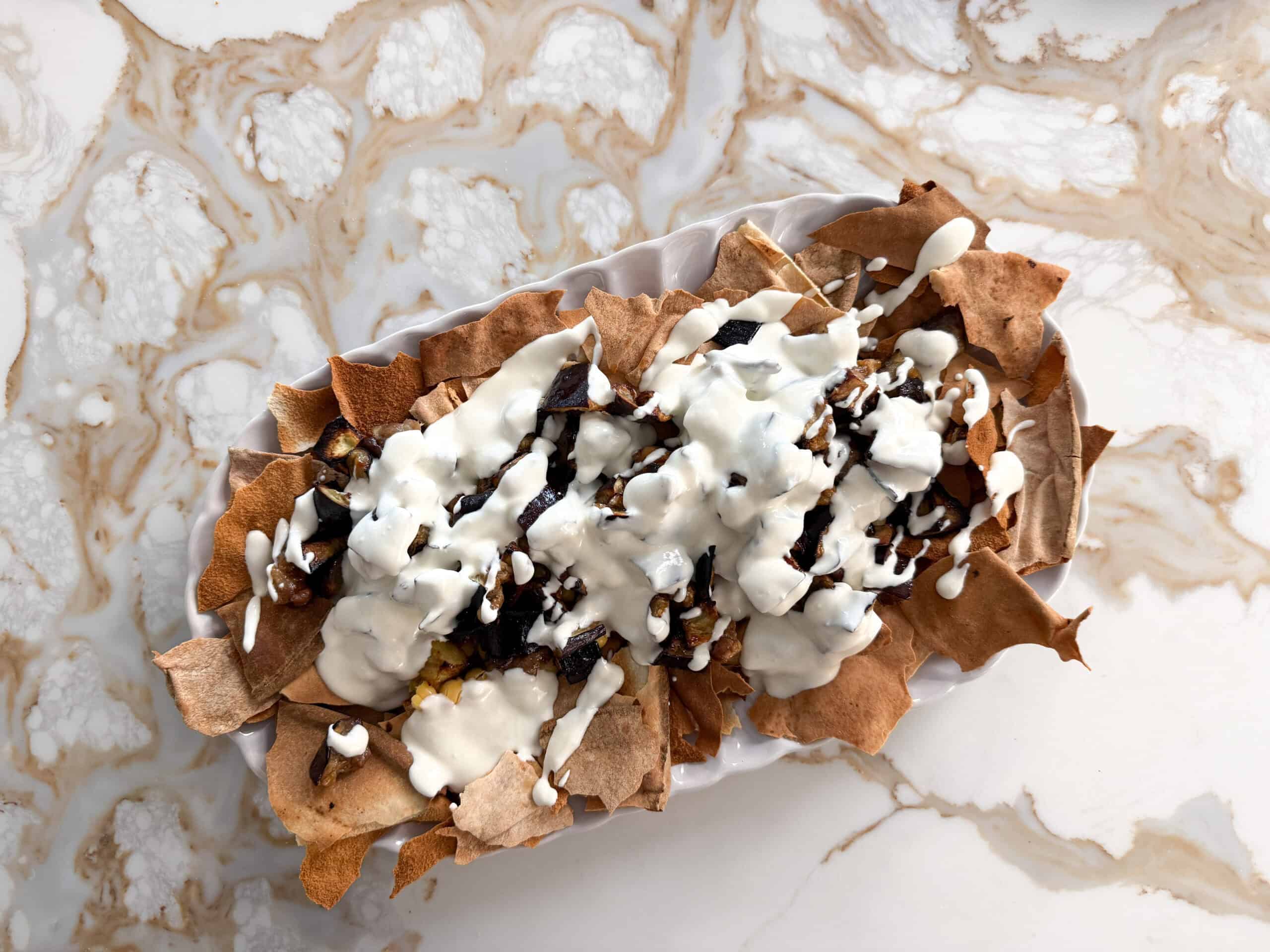 crispy broken pita bread with chickpeas, roasted eggplant pieces and white yogurt sauce on a white plate